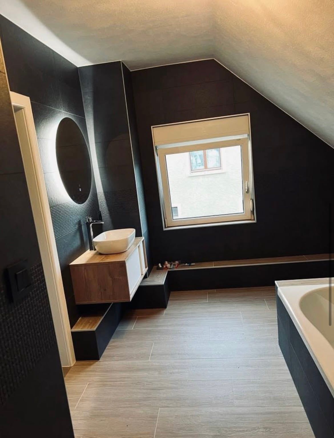 Salle de bains sous combles : carrelage sombre, vasque posée sur un meuble en bois, miroir rond éclairé, baignoire encastrée